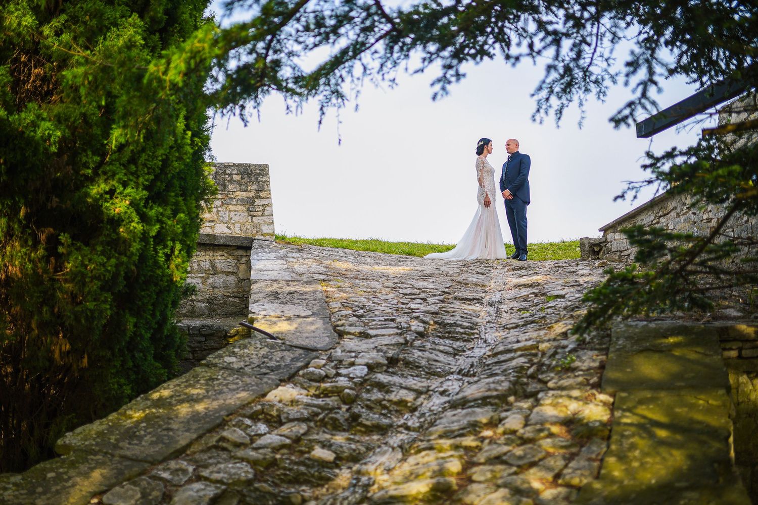 Bride and groom sharing a quiet wedding portrait on a historic stone path surrounded by trees and old architecture. By Herzenshaus Studio.