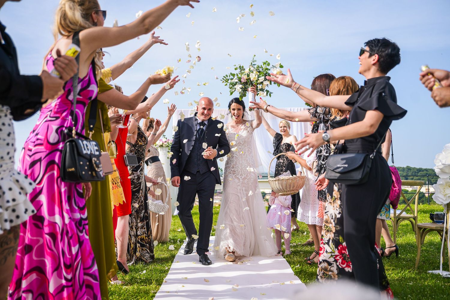Bride and groom walk down outdoor aisle as guests toss colorful confetti at sunny wedding celebration.