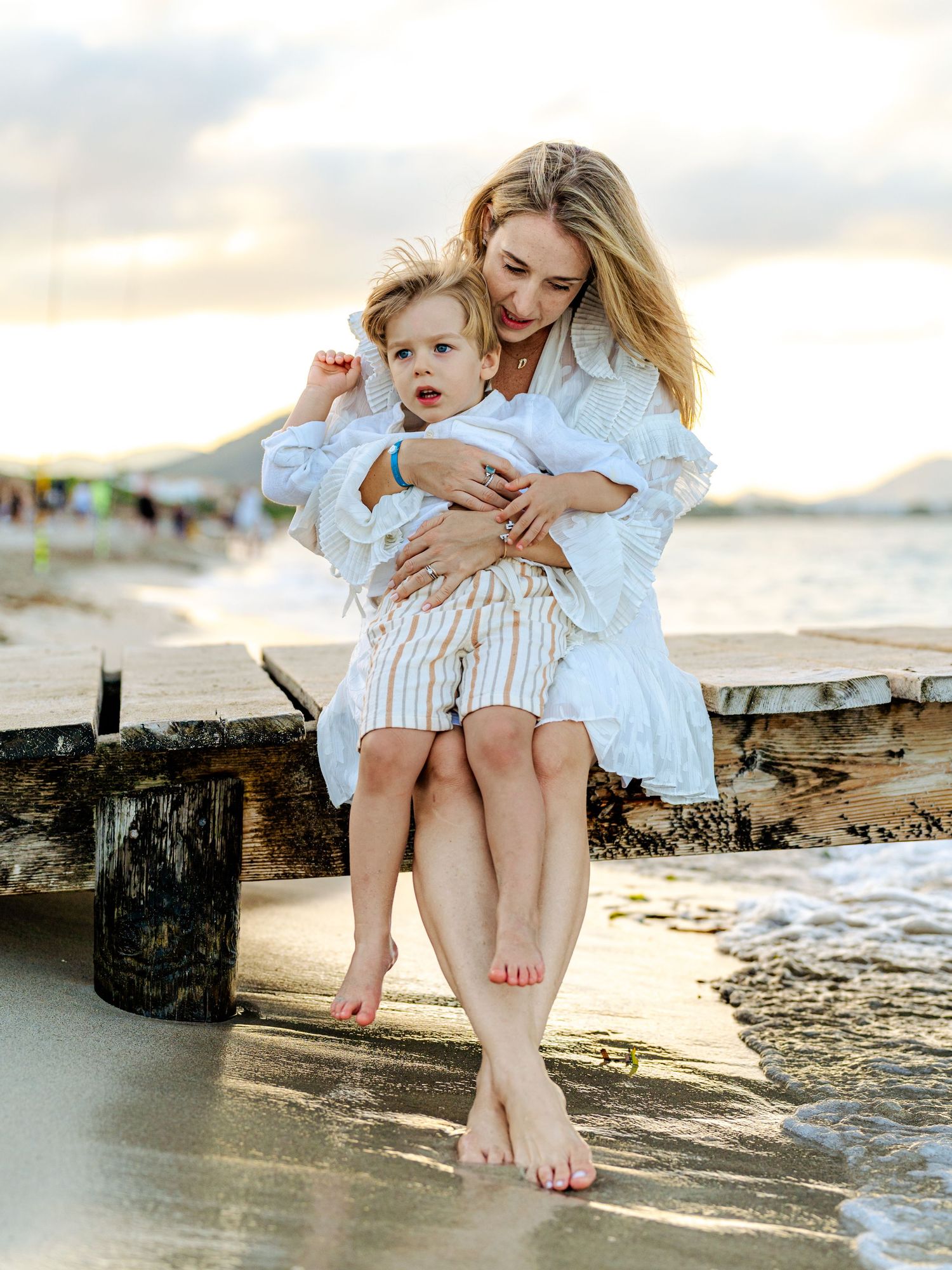 Mother sitting on beach pier holding toddler son at sunset.