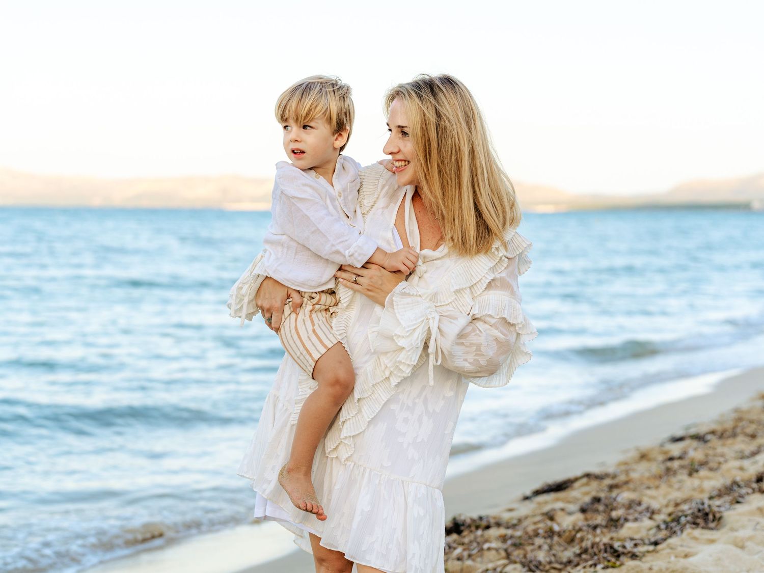Smiling blonde mother holding toddler son on sunny beach.