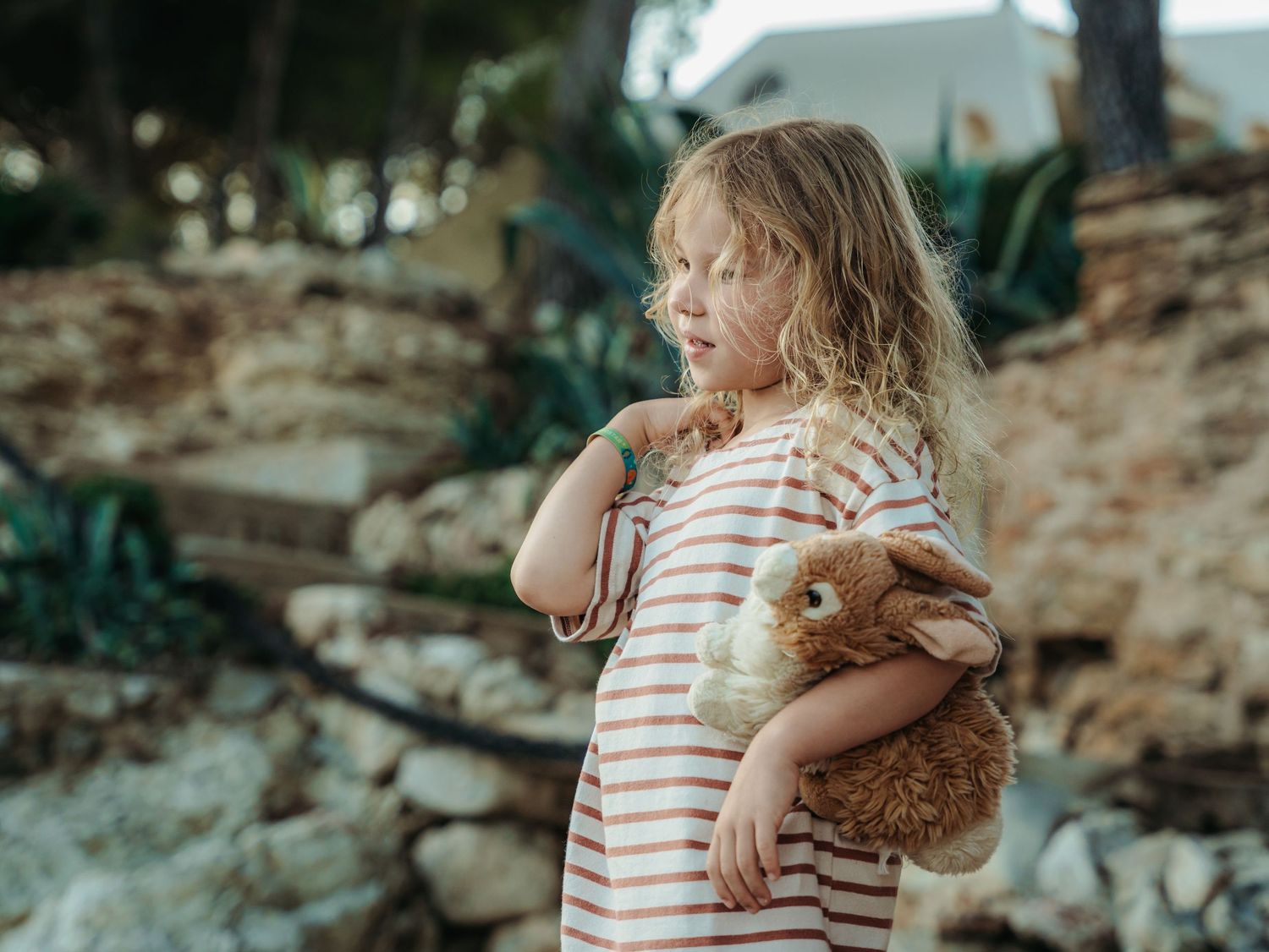 A young girl in a striped dress holds a fluffy bunny stuffed animal in a rocky outdoor setting.