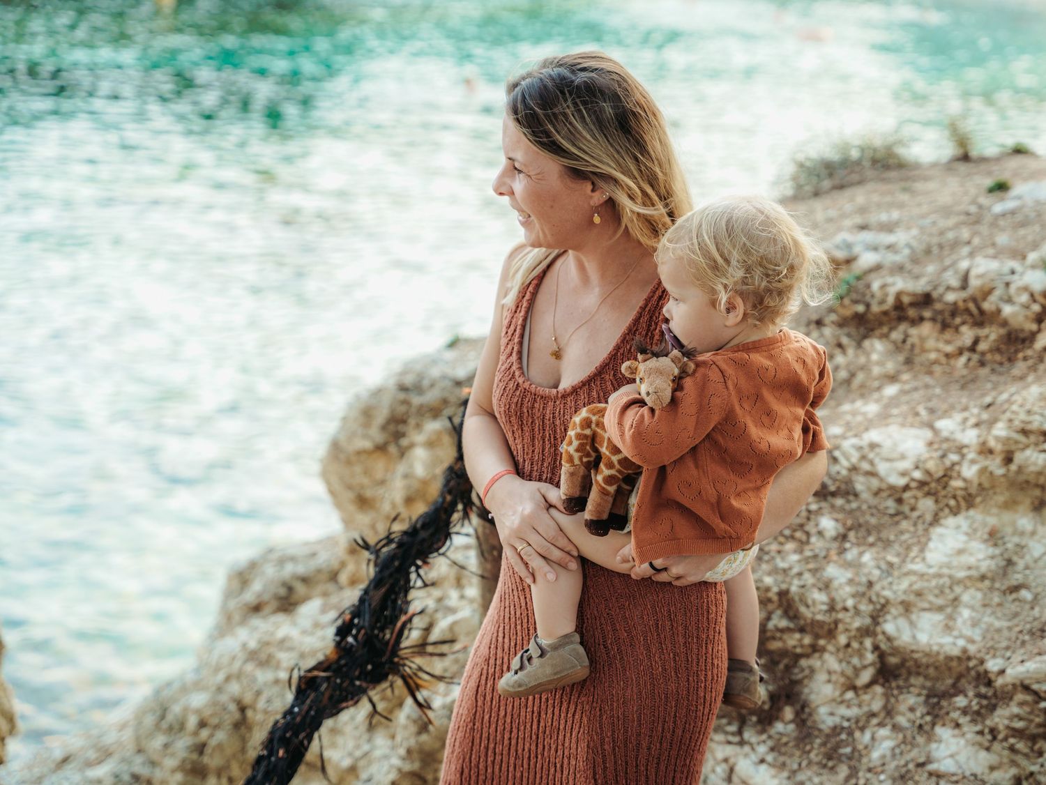 Woman in terracotta knit dress carrying toddler with stuffed giraffe on sun-drenched rocky coastline.