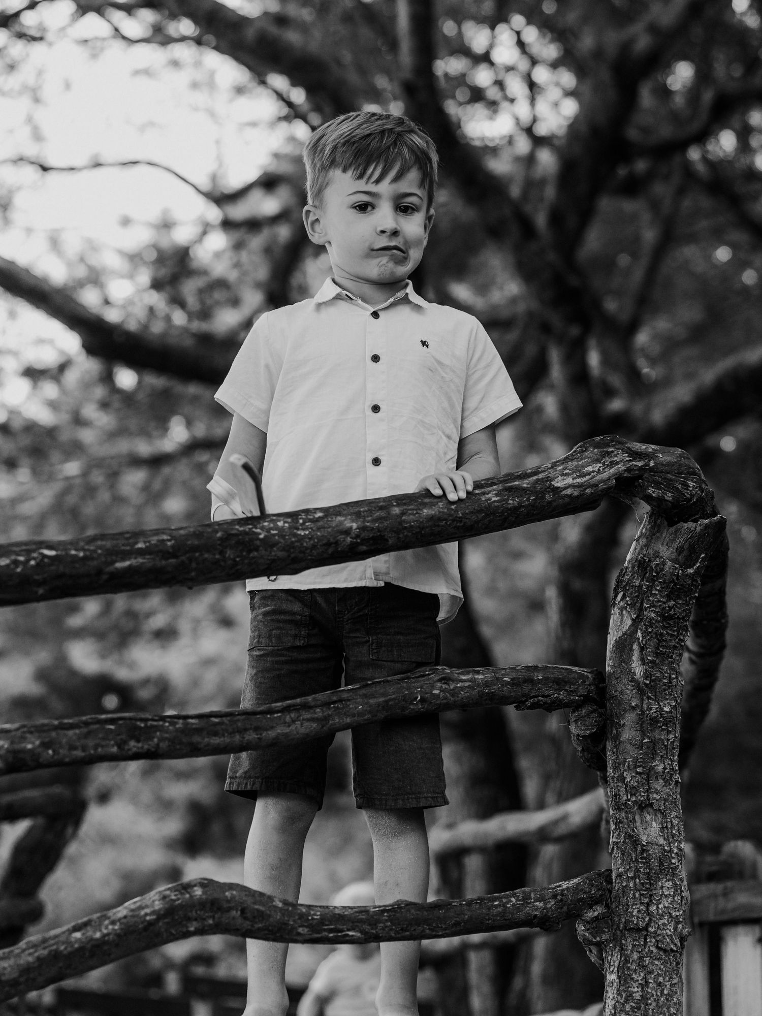 Black and white portrait of a young boy in white shirt and shorts standing on a rustic wooden fence outdoors.