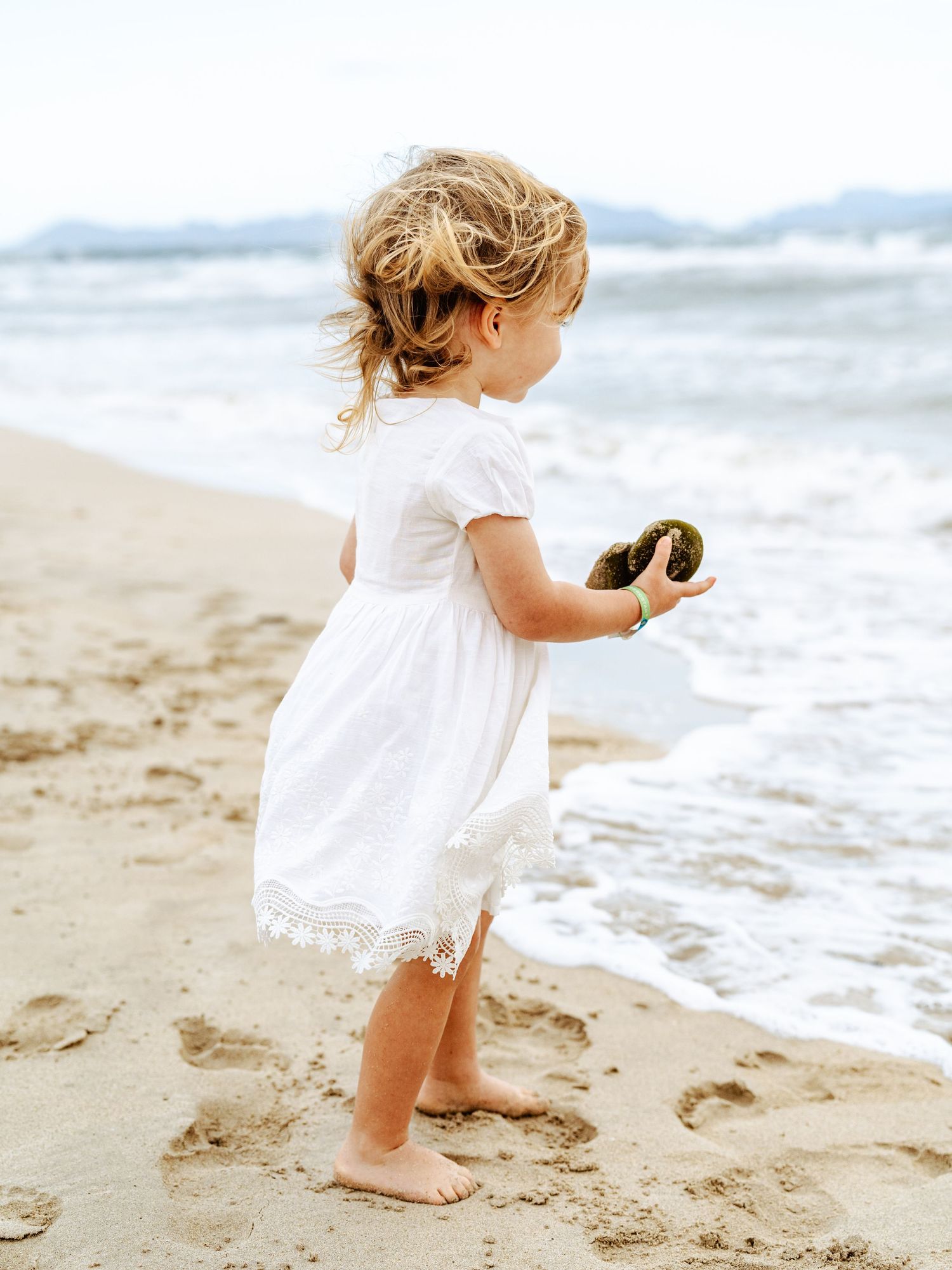Young girl in white dress standing barefoot on beach sand holding a rock near ocean waves.
