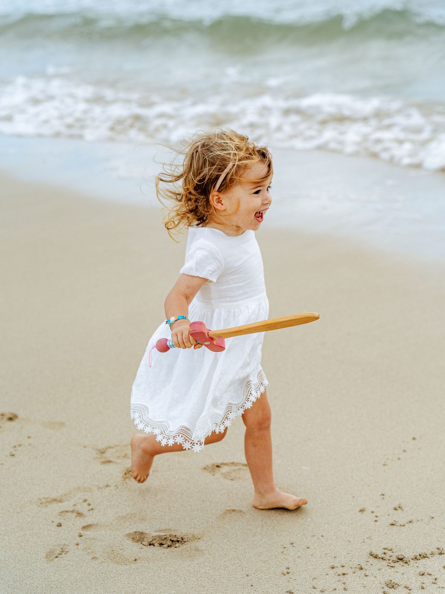 Cute toddler girl in white dress holding a wooden toy while walking on the beach.