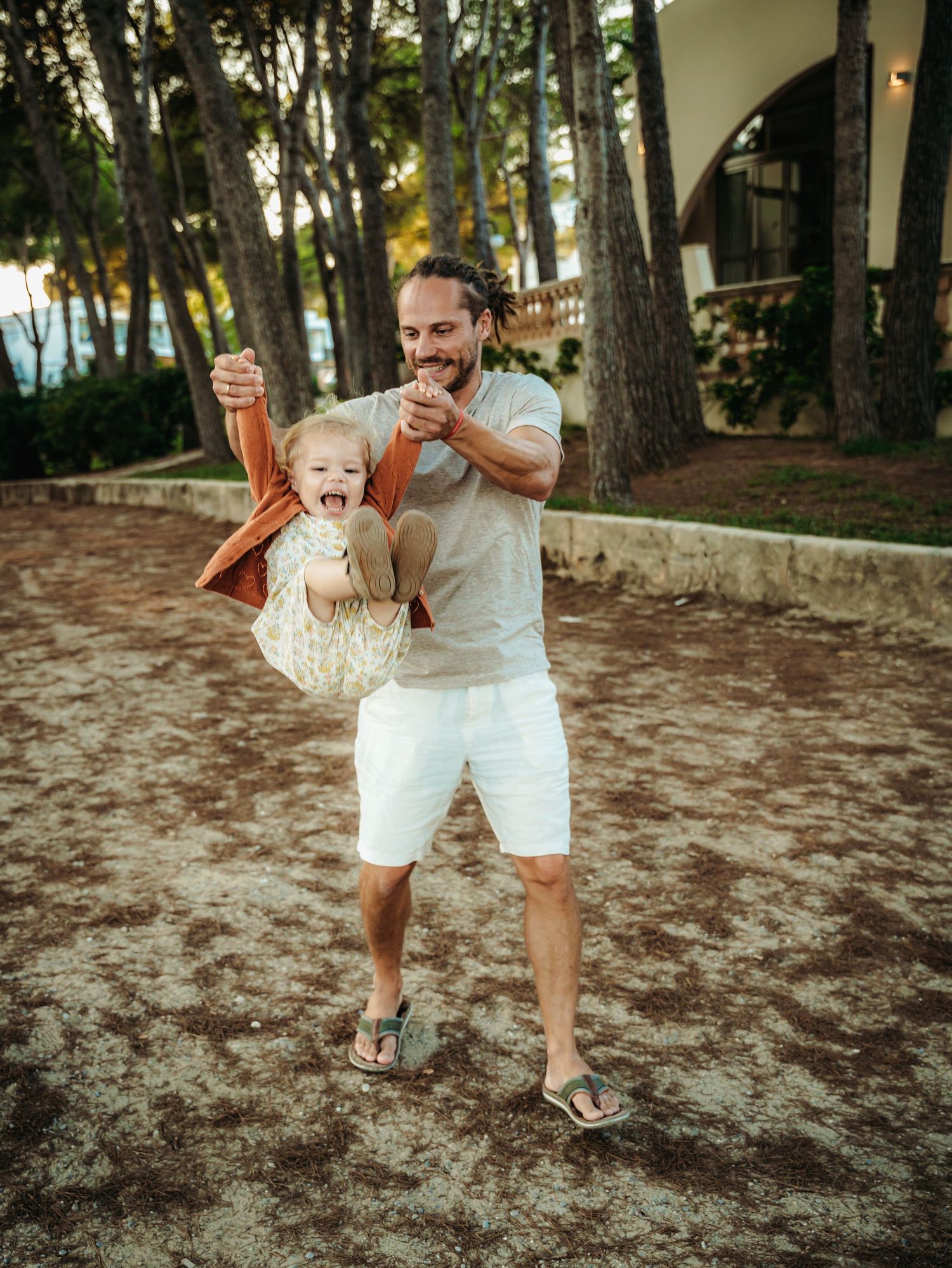 Father joyfully lifting laughing toddler in rust cardigan outdoors among tall pine trees.
