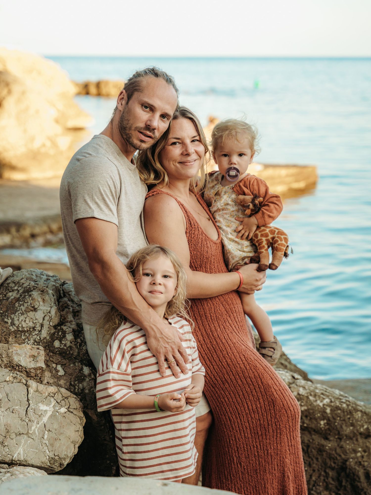 Family of four poses on rocky seaside, pregnant mother holding baby while father embraces young child.