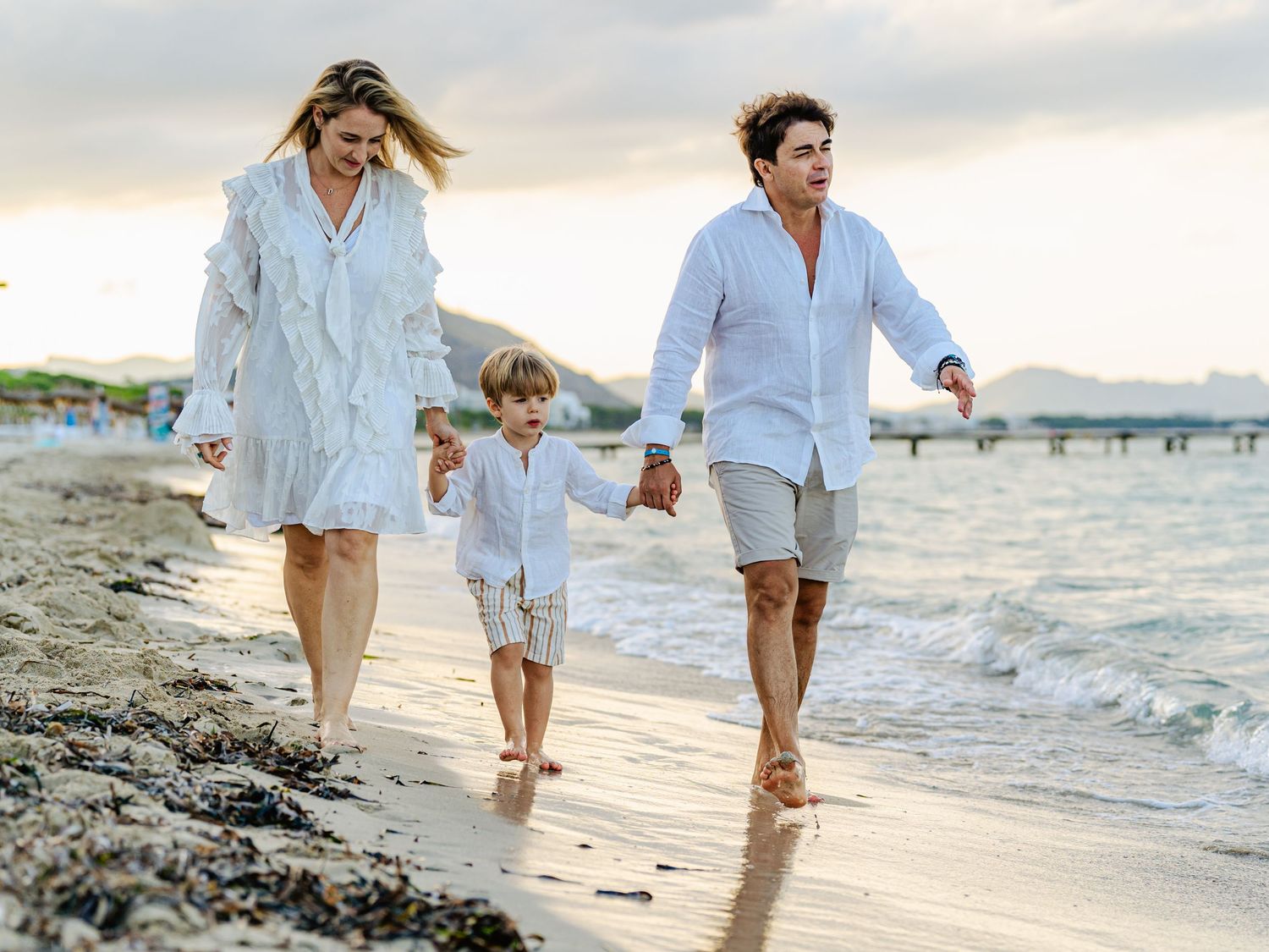 Smiling family with young child walking hand in hand along a scenic beach at golden sunset hour.