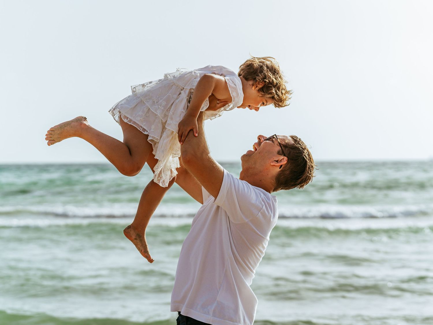 Father lifts laughing daughter in white dress high above beach with ocean in background.