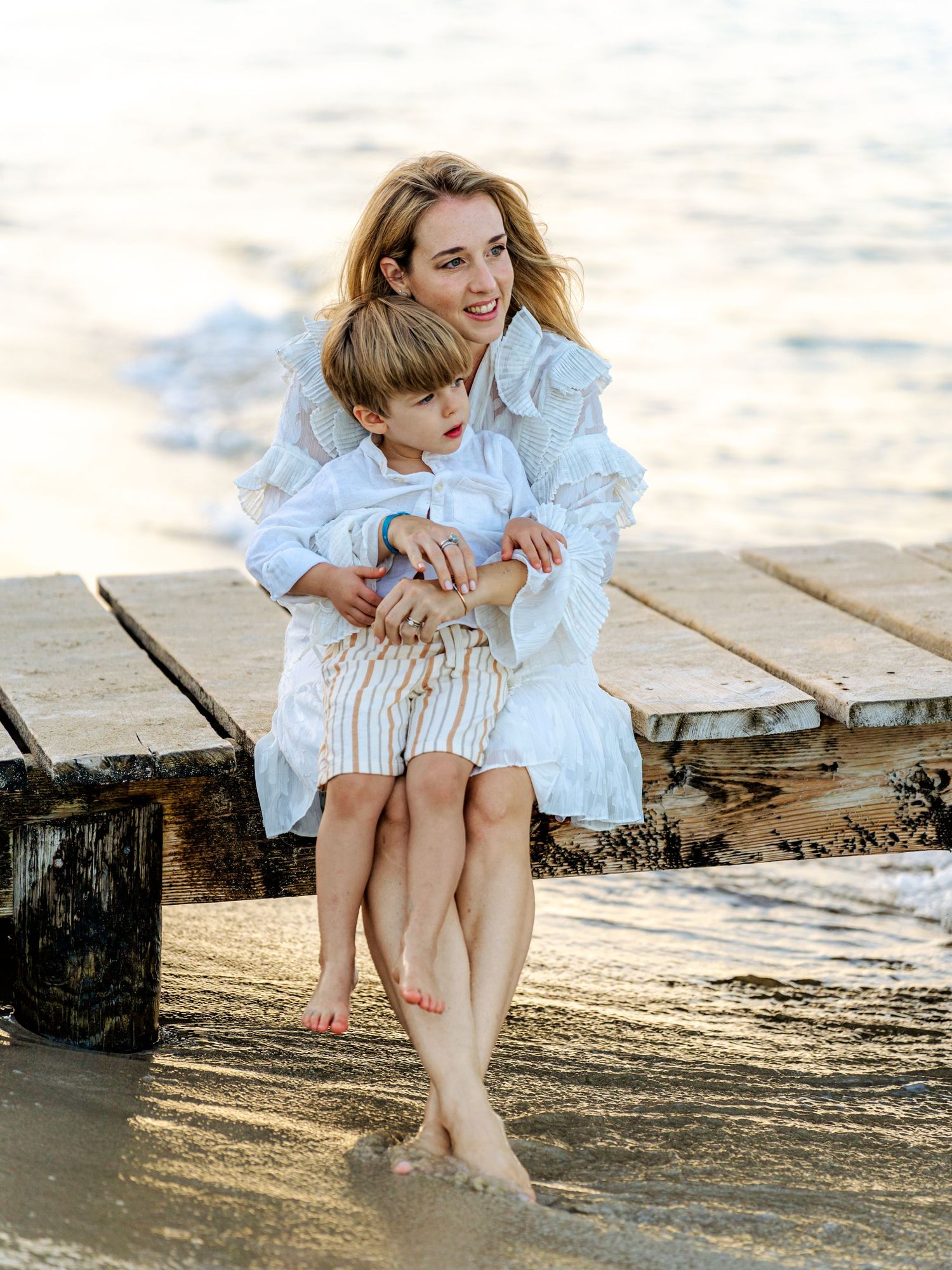 A mother in a white dress sitting on a beach dock cuddling her young son at sunset.