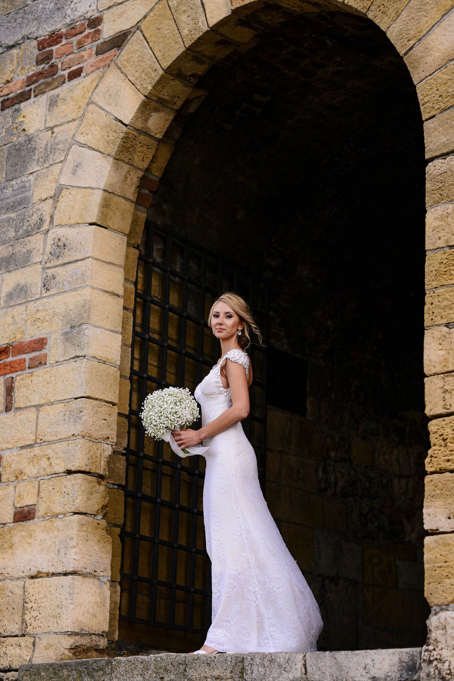 Elegant blonde bride in fitted lace gown holding white bouquet stands in a historic stone arched doorway.