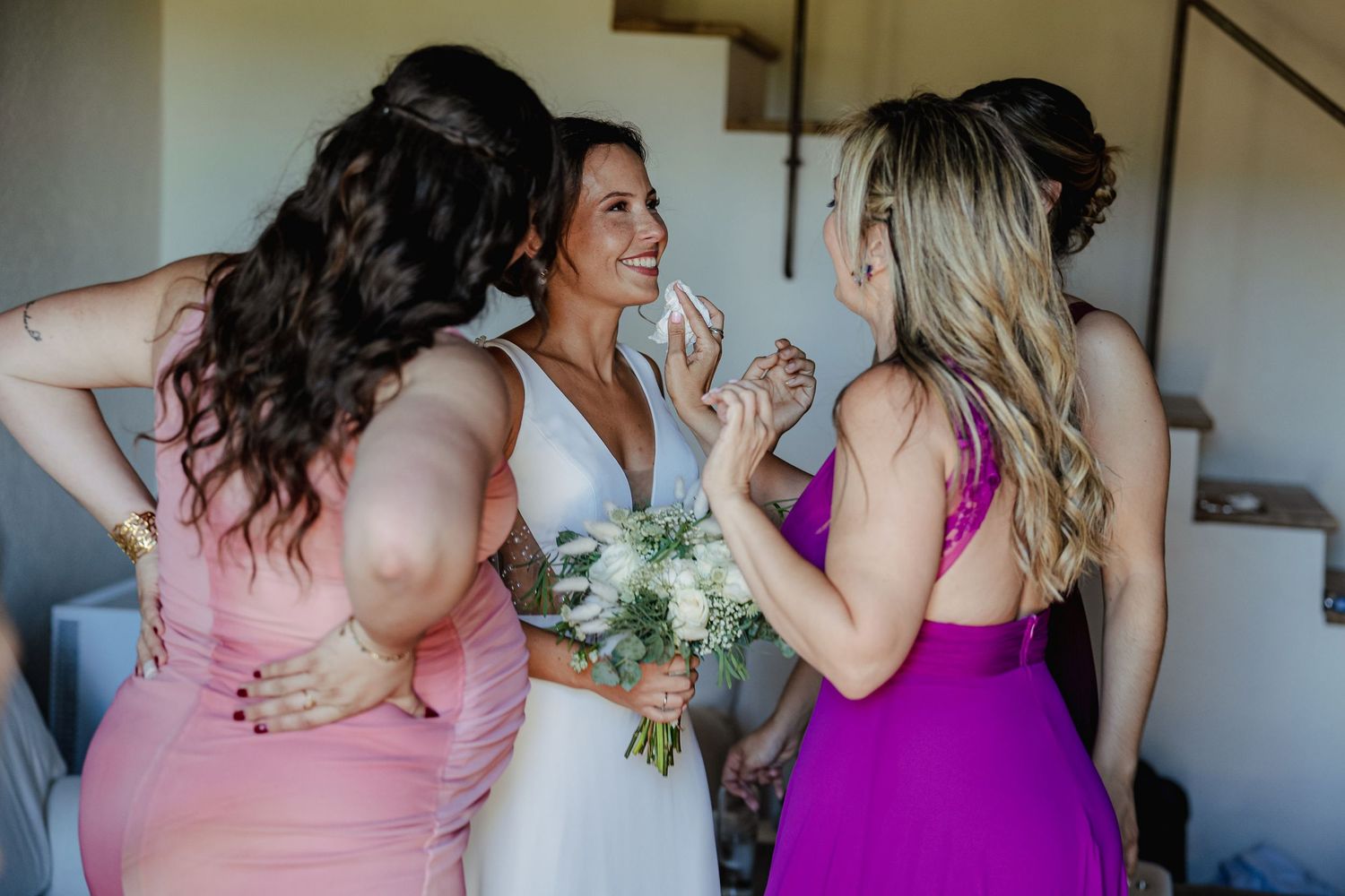 Bride holding white bouquet sharing a joyful moment with bridesmaids in pink and purple dresses.