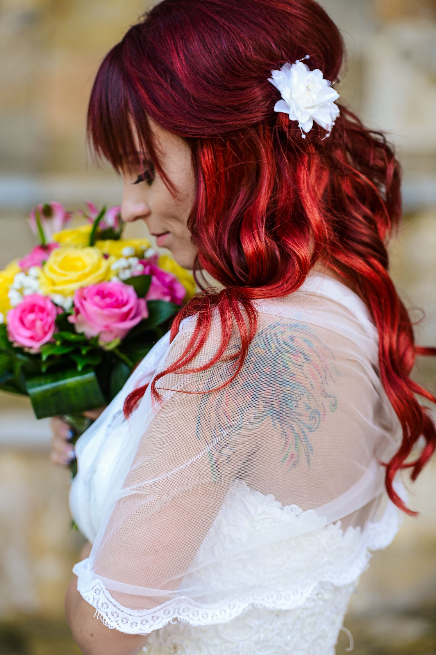 Red-haired bride with shoulder tattoo holding colorful bouquet of pink and yellow roses in white lace dress.
