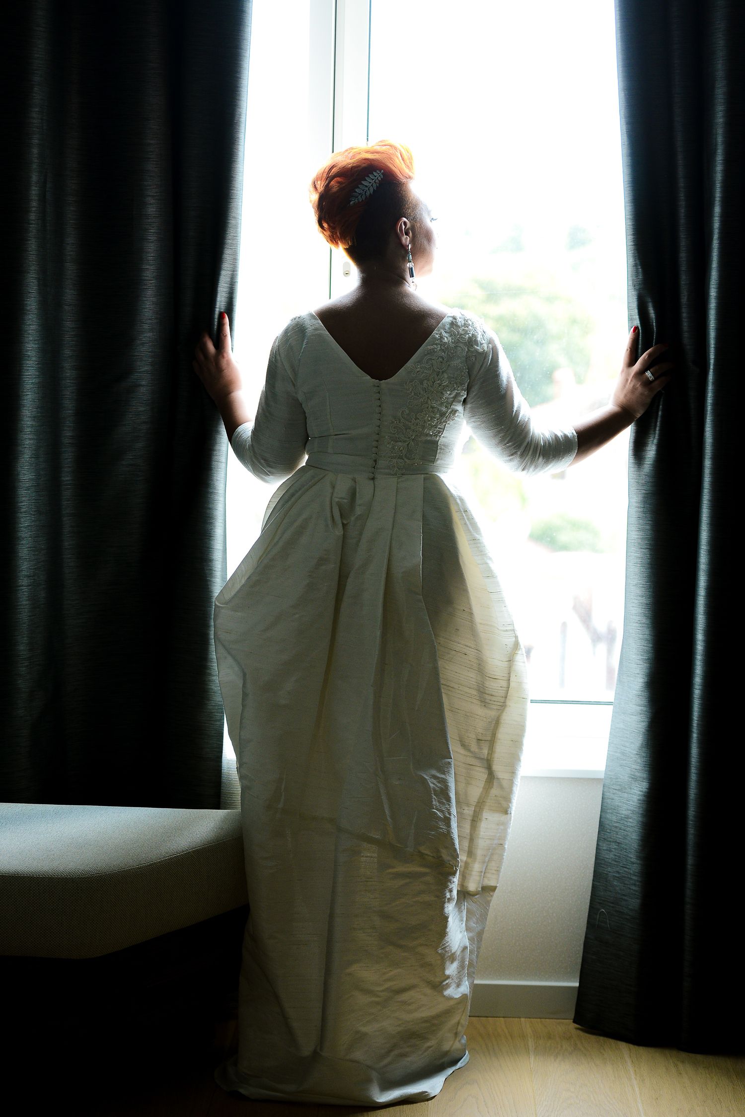 Elegant bride in a long-sleeve white wedding gown standing at a window with dark curtains.