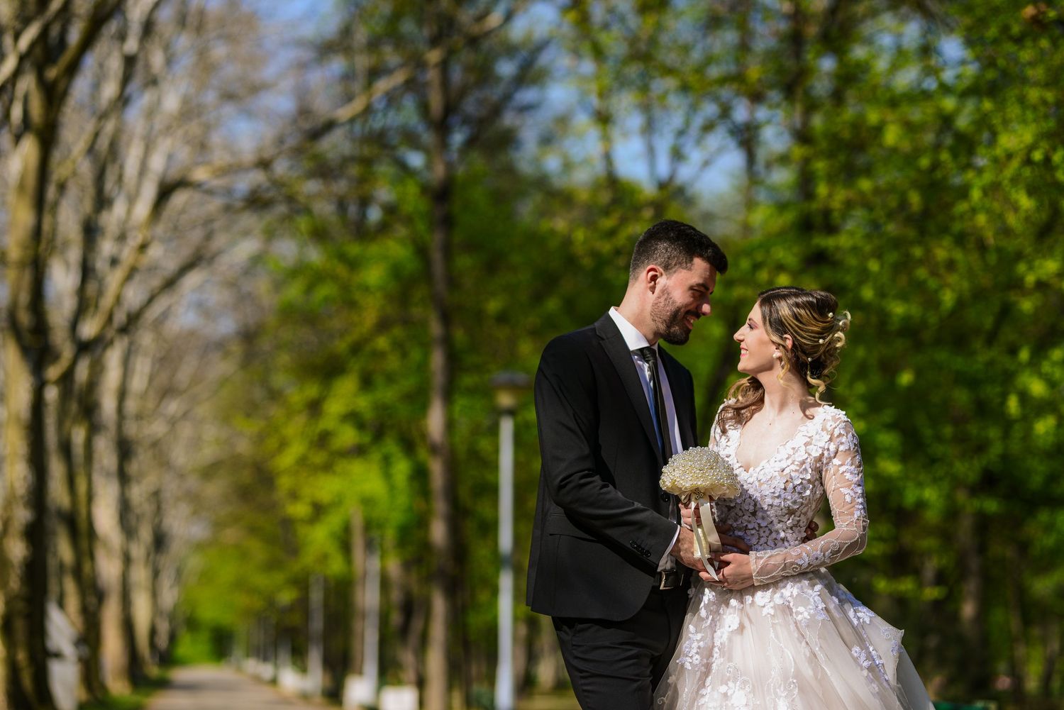 Bride and groom share a romantic gaze while walking through a tree-lined path on their wedding day.