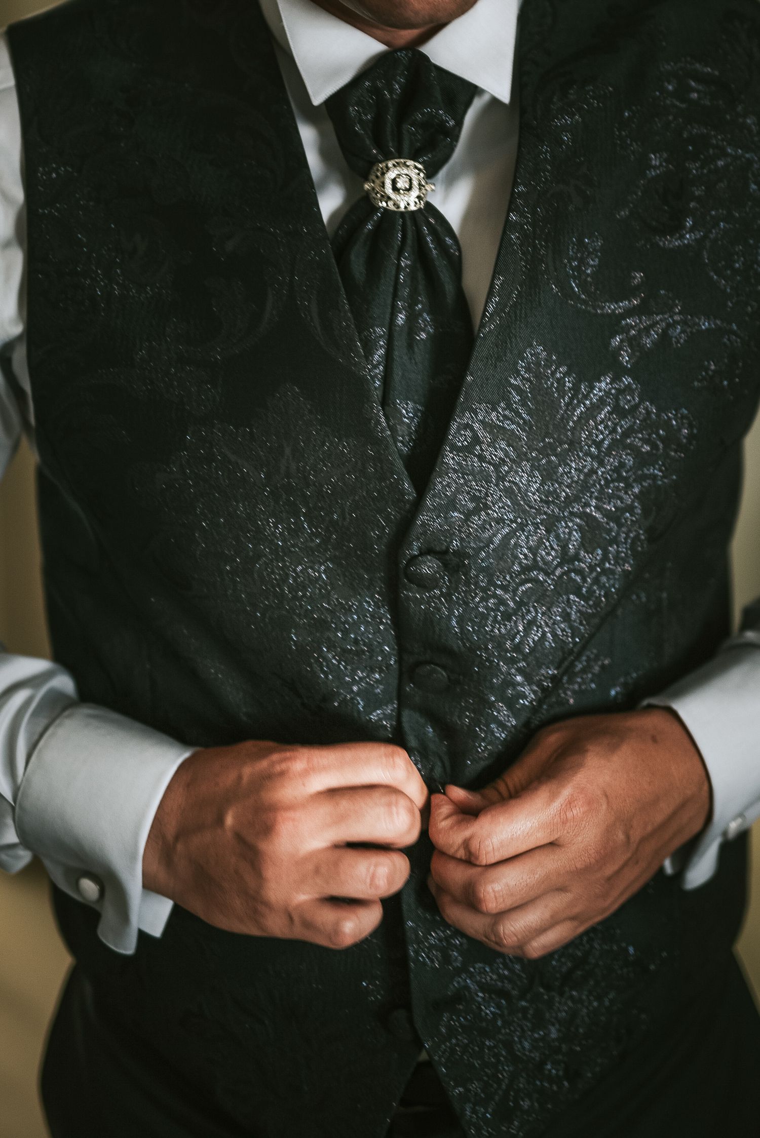 Groom buttoning an ornate dark floral patterned vest with a decorative brooch on his wedding day.