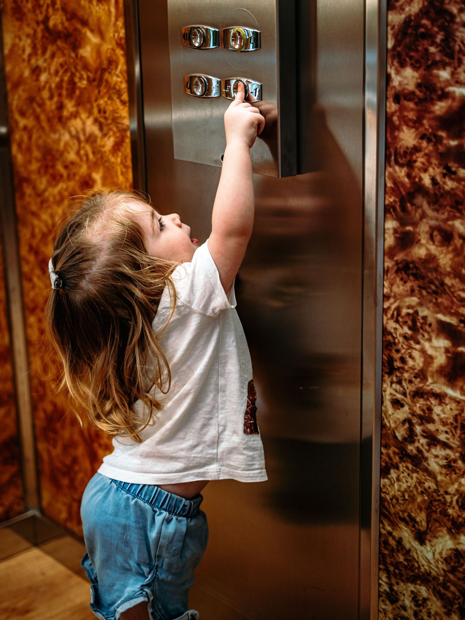 Young toddler girl in white shirt reaching up to press elevator buttons indoors.