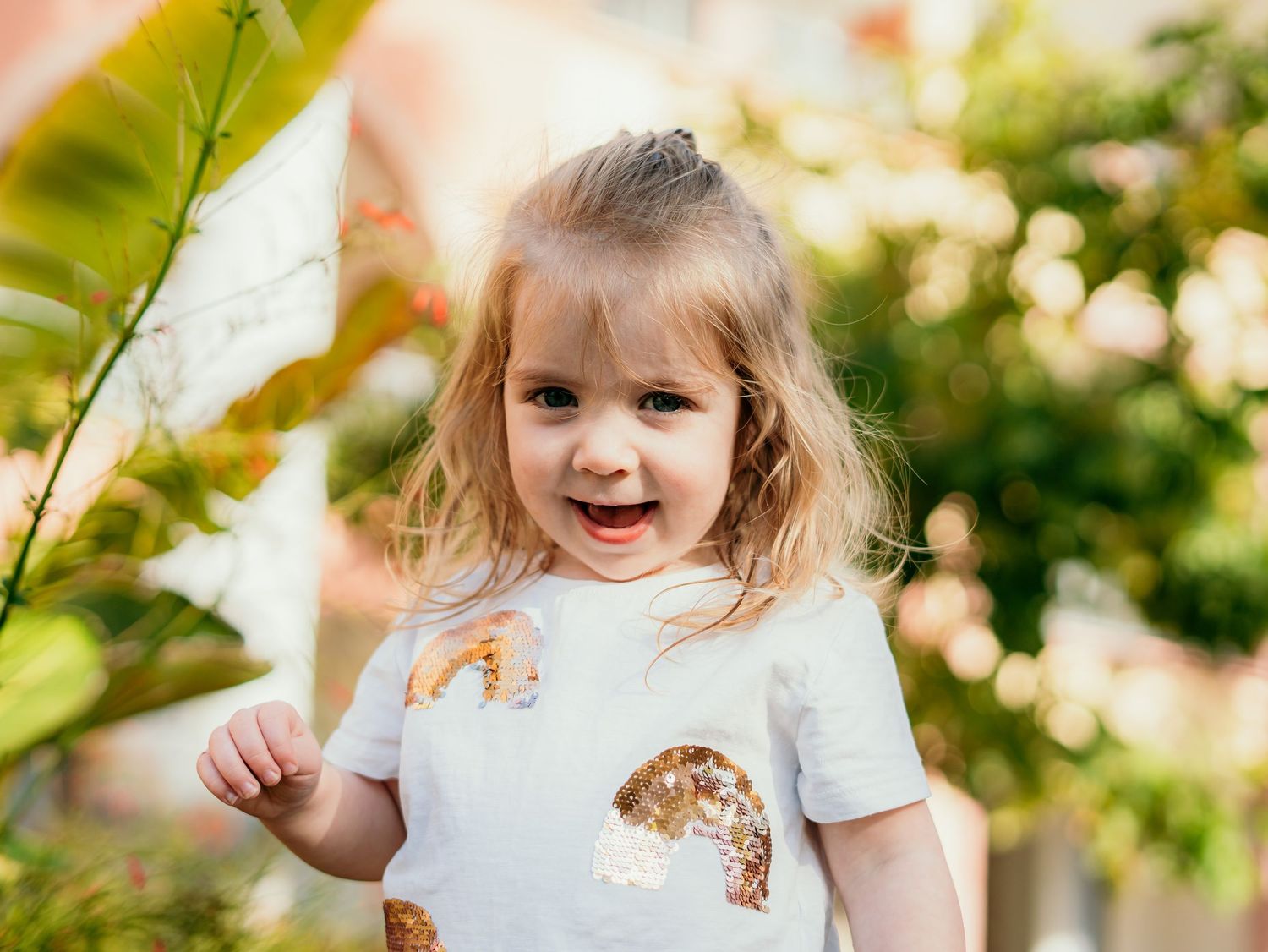 Happy blonde toddler girl in a sequin rainbow shirt laughing joyfully in a sunny garden.