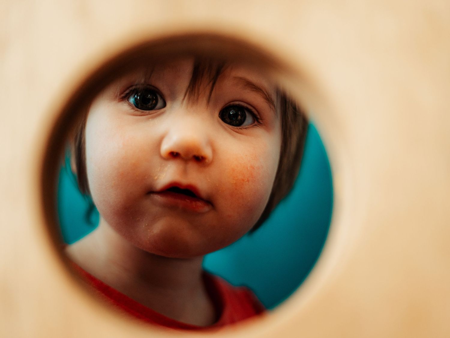 Close-up of toddler girl with big brown eyes peeking through circular hole in wooden toy.
