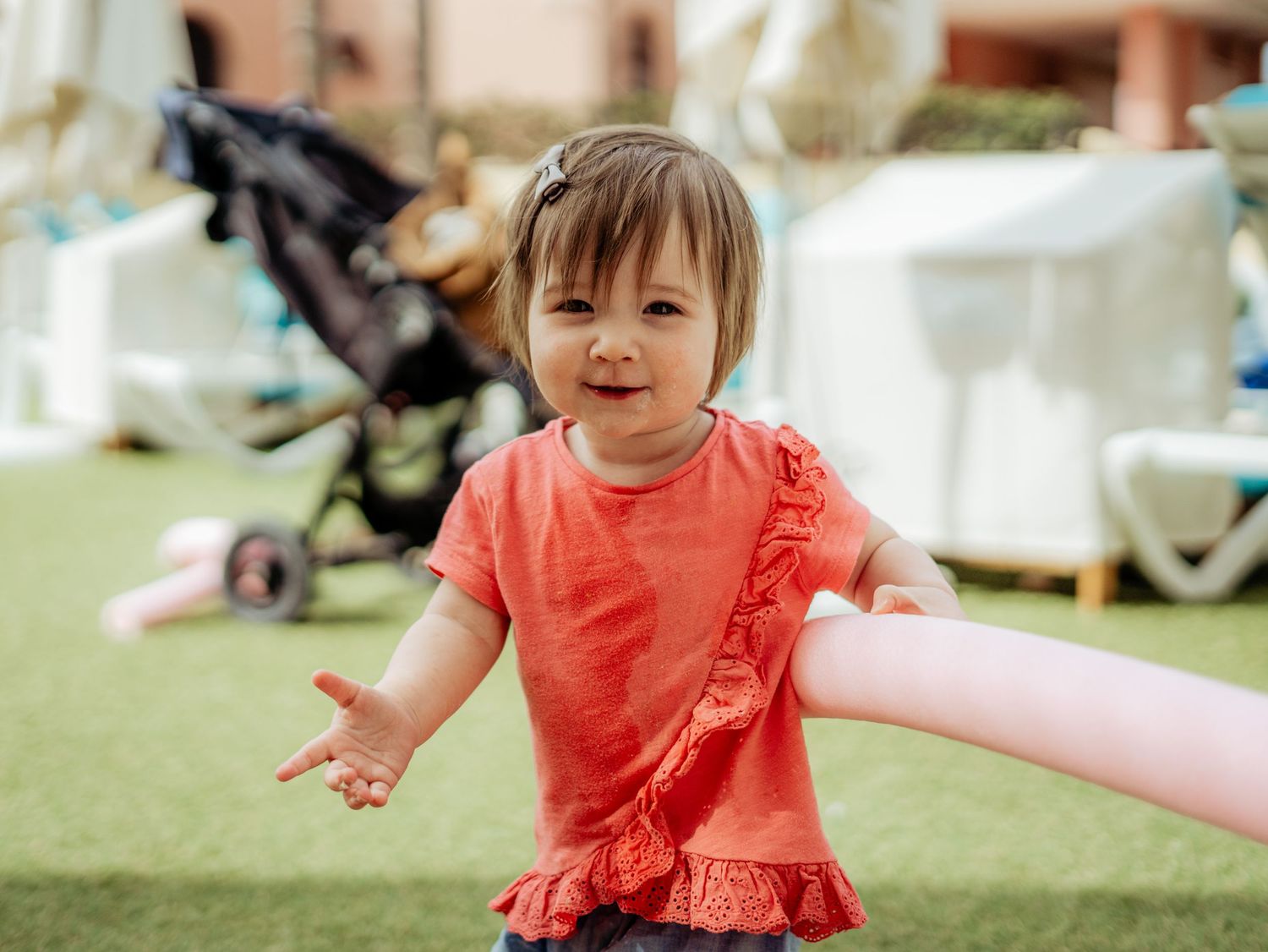 Joyful toddler girl in red ruffled top reaching out arms on a green lawn outdoors.