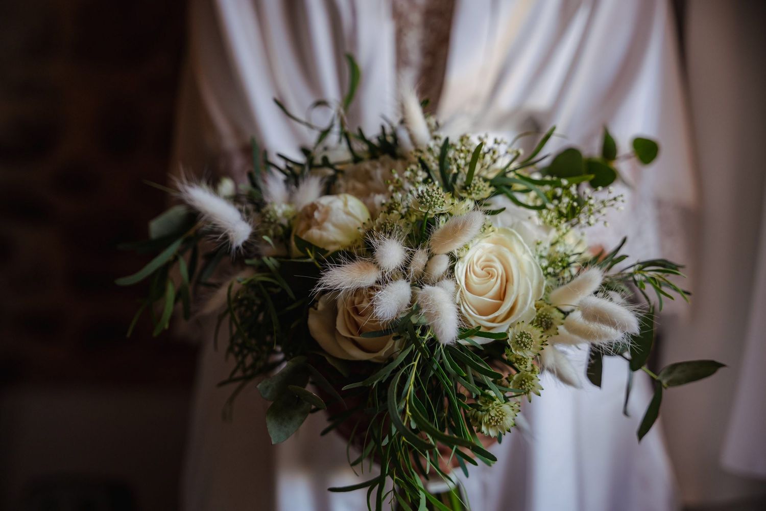 Bride in white robe holding elegant bouquet of white roses and pampas grass.