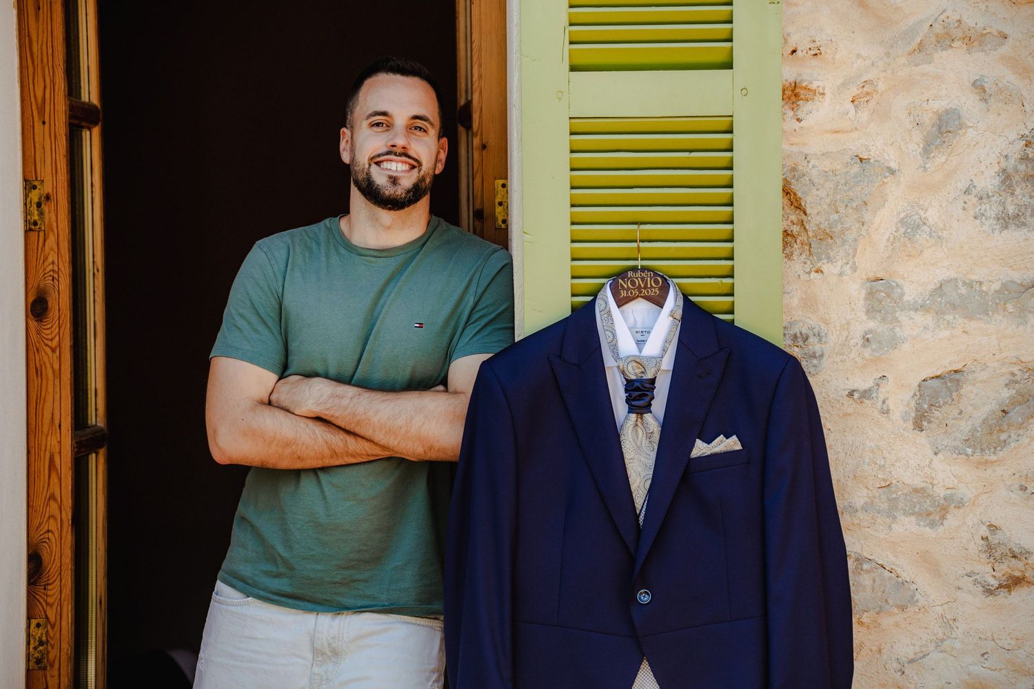 Groom smiling beside his navy blue wedding suit hanging outdoors against a stone wall.