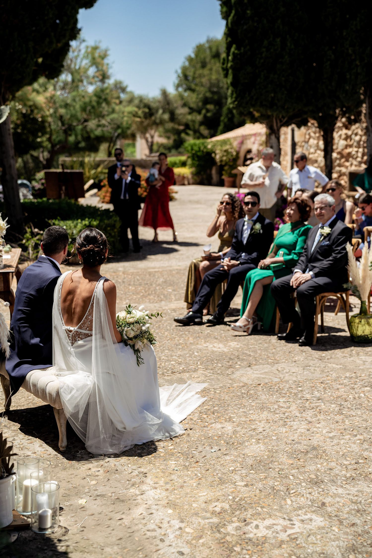 Outdoor wedding ceremony with bride and groom seated as guests watch in sunlit garden setting.