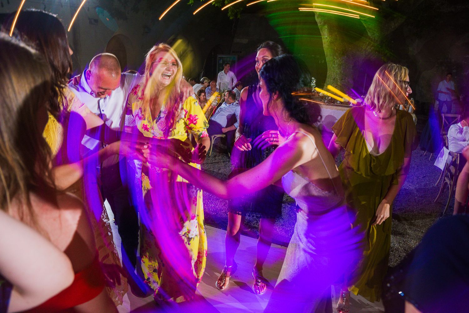 Wedding guests dance energetically under colorful purple lights at an outdoor nighttime reception party.