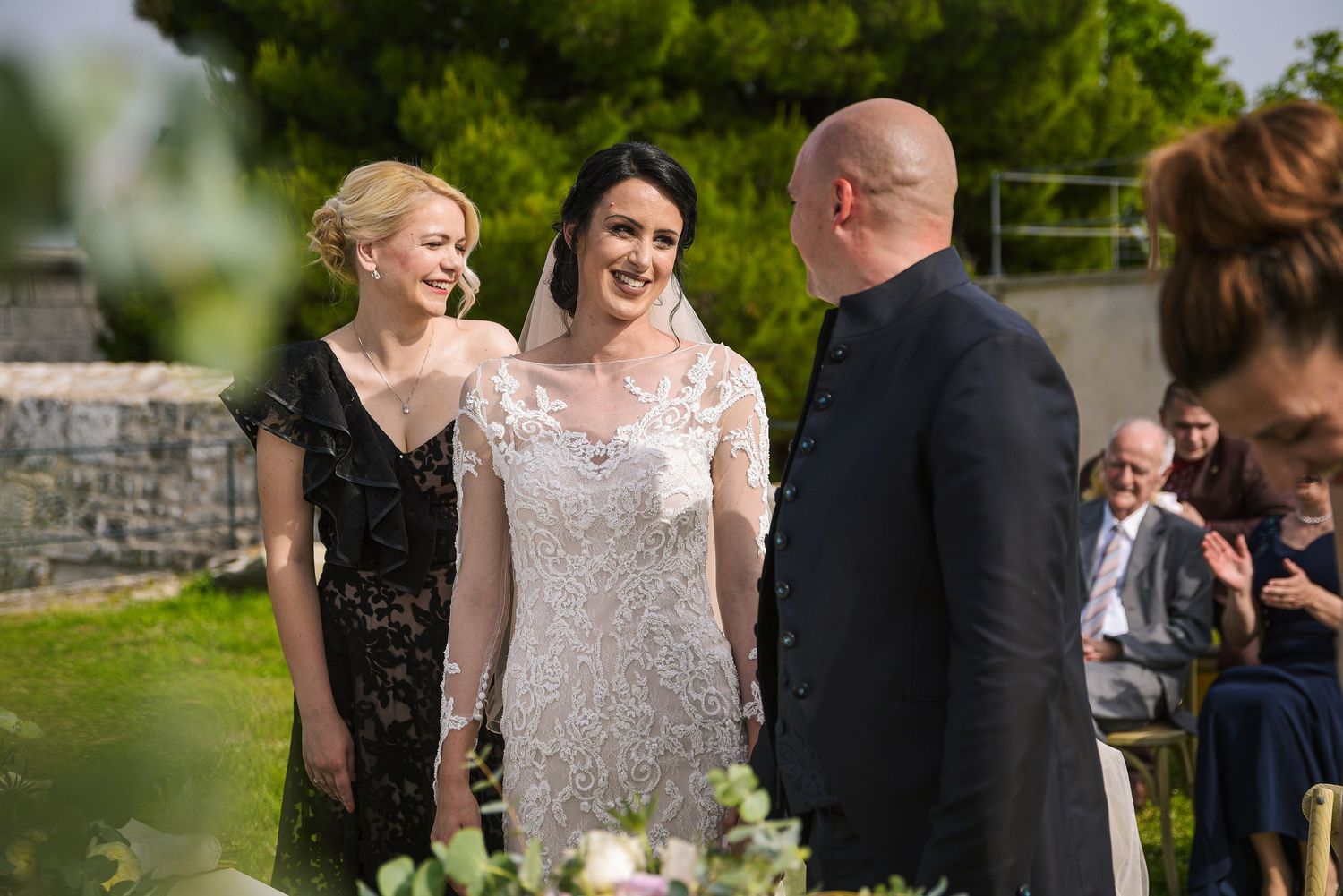 Bride in lace gown smiles at groom during outdoor wedding ceremony with guests seated.