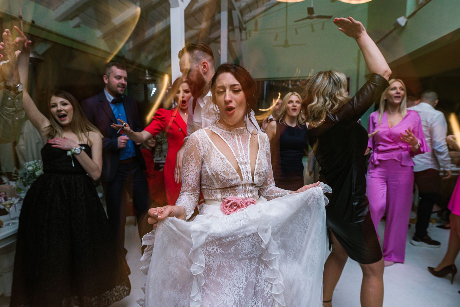 Bride in lace gown lifts her dress while dancing energetically with wedding guests.