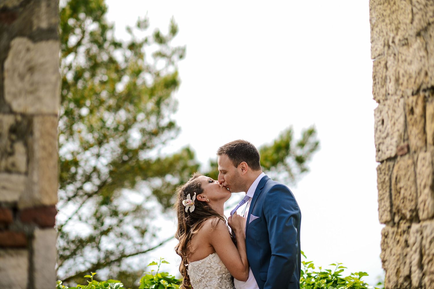 Wedding couple sharing a kiss between stone columns with lush green trees in background.