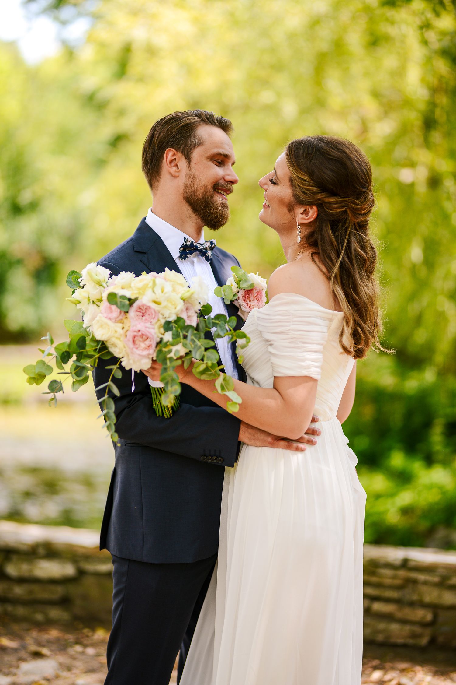 Bearded groom in navy tuxedo holds bride close outdoors, both smiling, with a lush floral bouquet.
