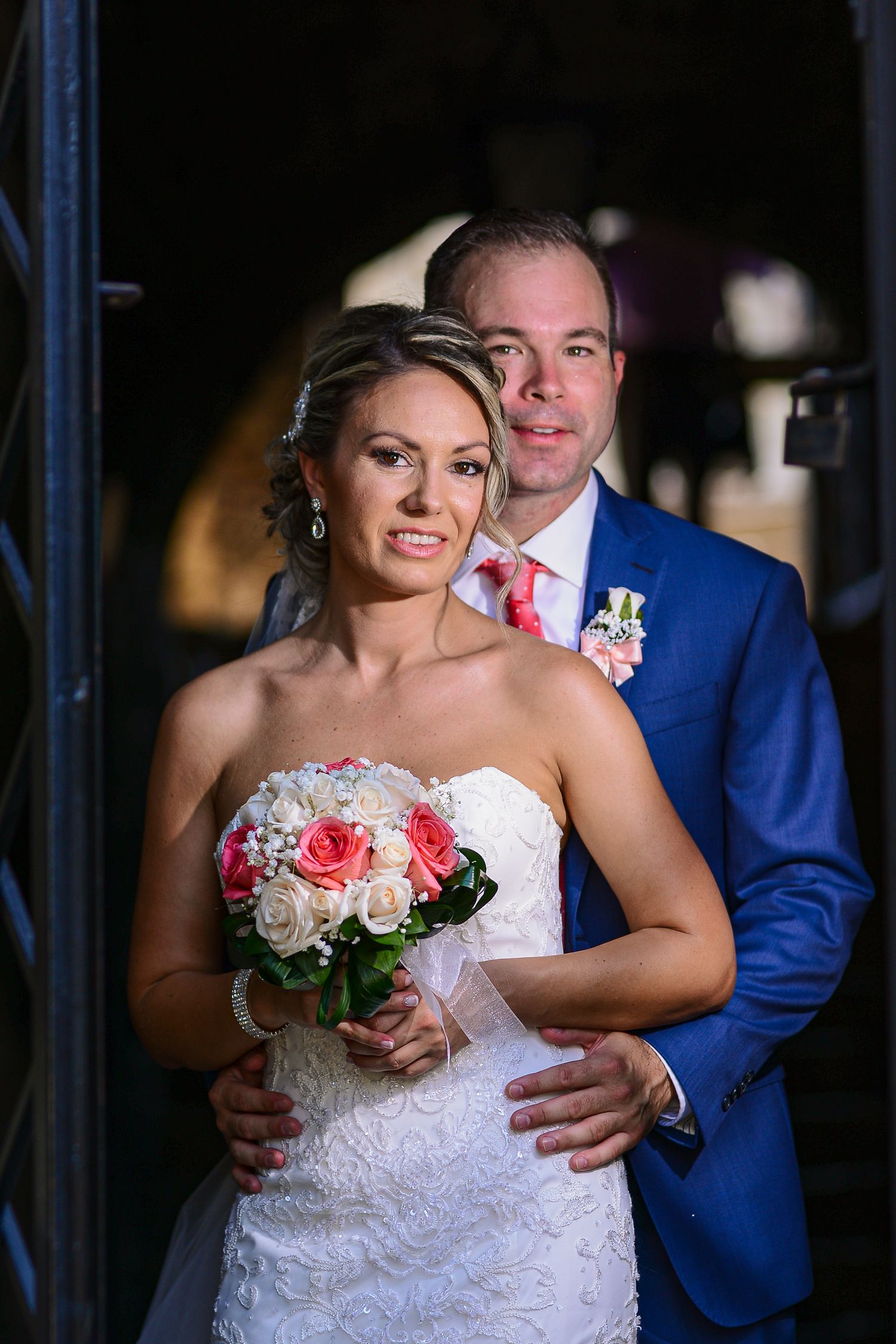 Bride holding coral rose bouquet with groom in blue suit embracing her at a wedding venue.