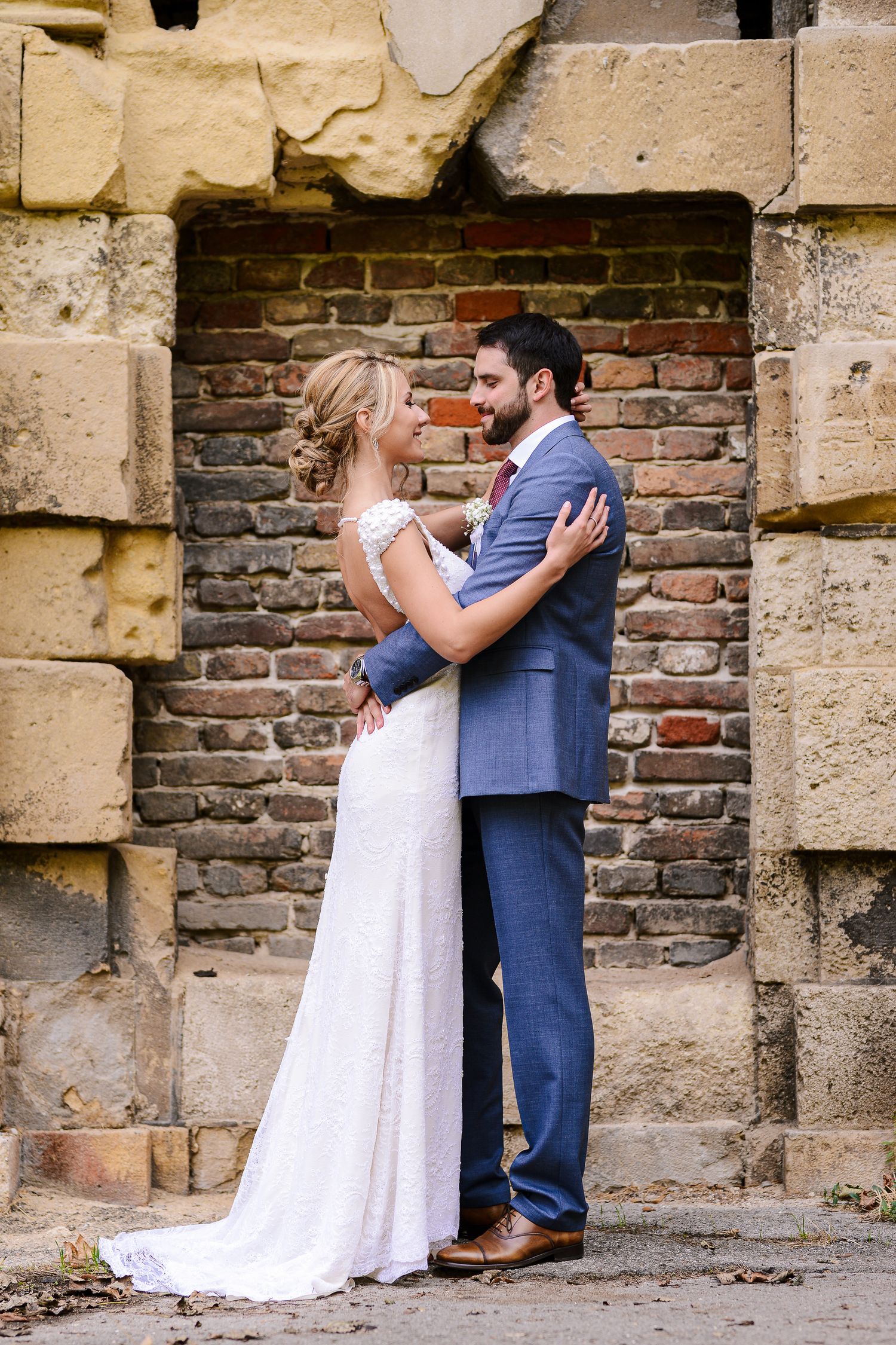 Bride and groom embrace lovingly in front of a rustic brick wall, she in lace gown, he in blue suit.