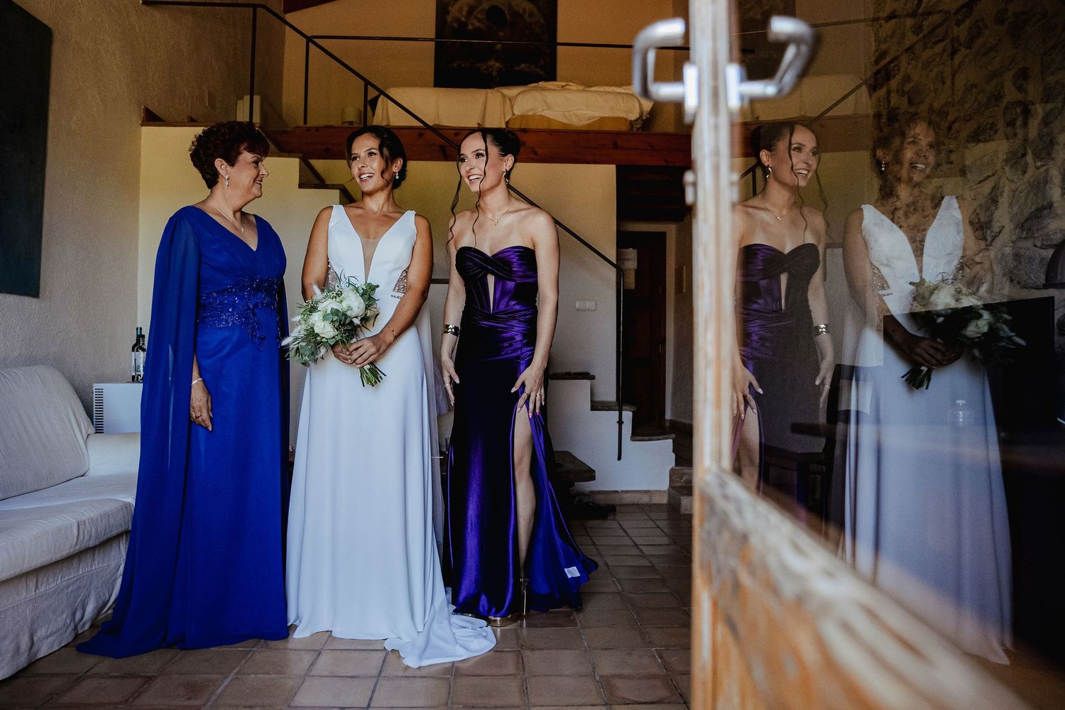 Bride with bridesmaids in royal blue and purple gowns standing together reflected in a glass door.