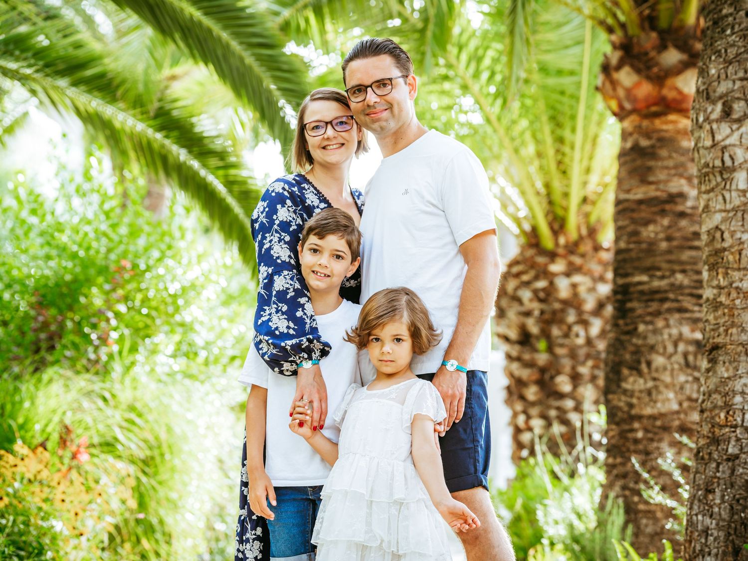 Family of four smiles together under palm trees in a lush tropical outdoor setting.