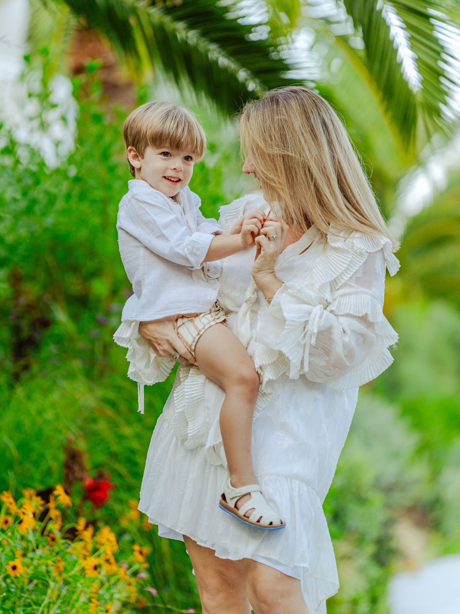 A smiling mother in a white ruffled dress holding her toddler son near tropical palm trees.