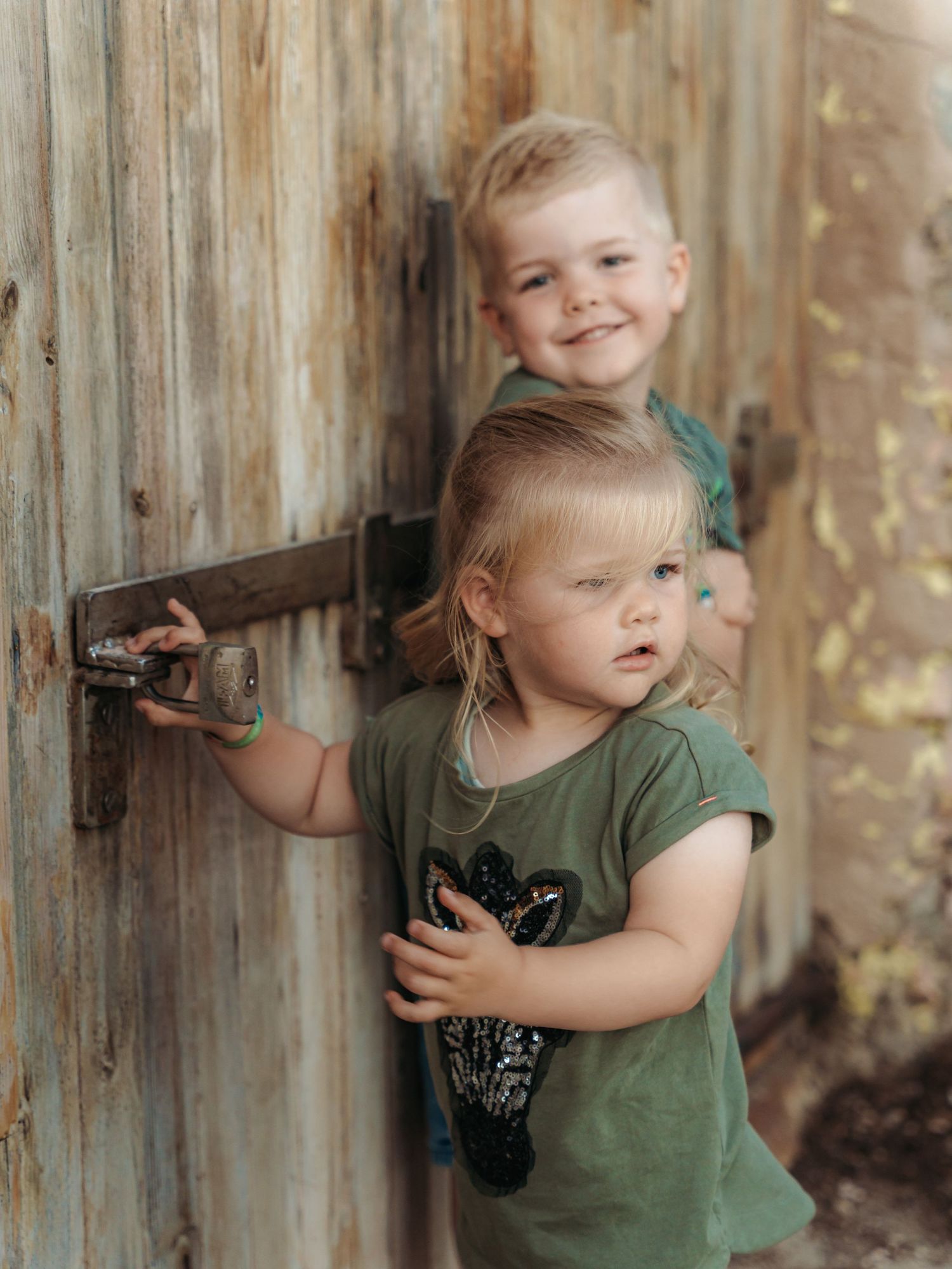 Two young siblings in matching green shirts peek curiously around a rustic wooden door.