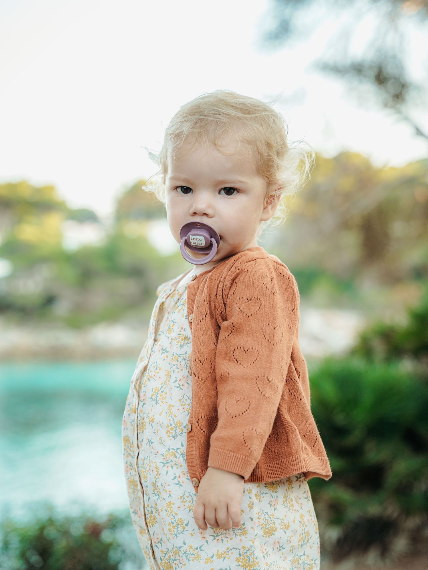 Blonde toddler girl with purple pacifier wearing floral dress and rust cardigan outdoors near water.