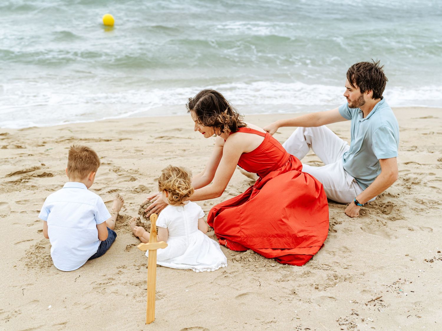 A family sits together on a sunny beach, with a mother in a red dress leaning over two young children.