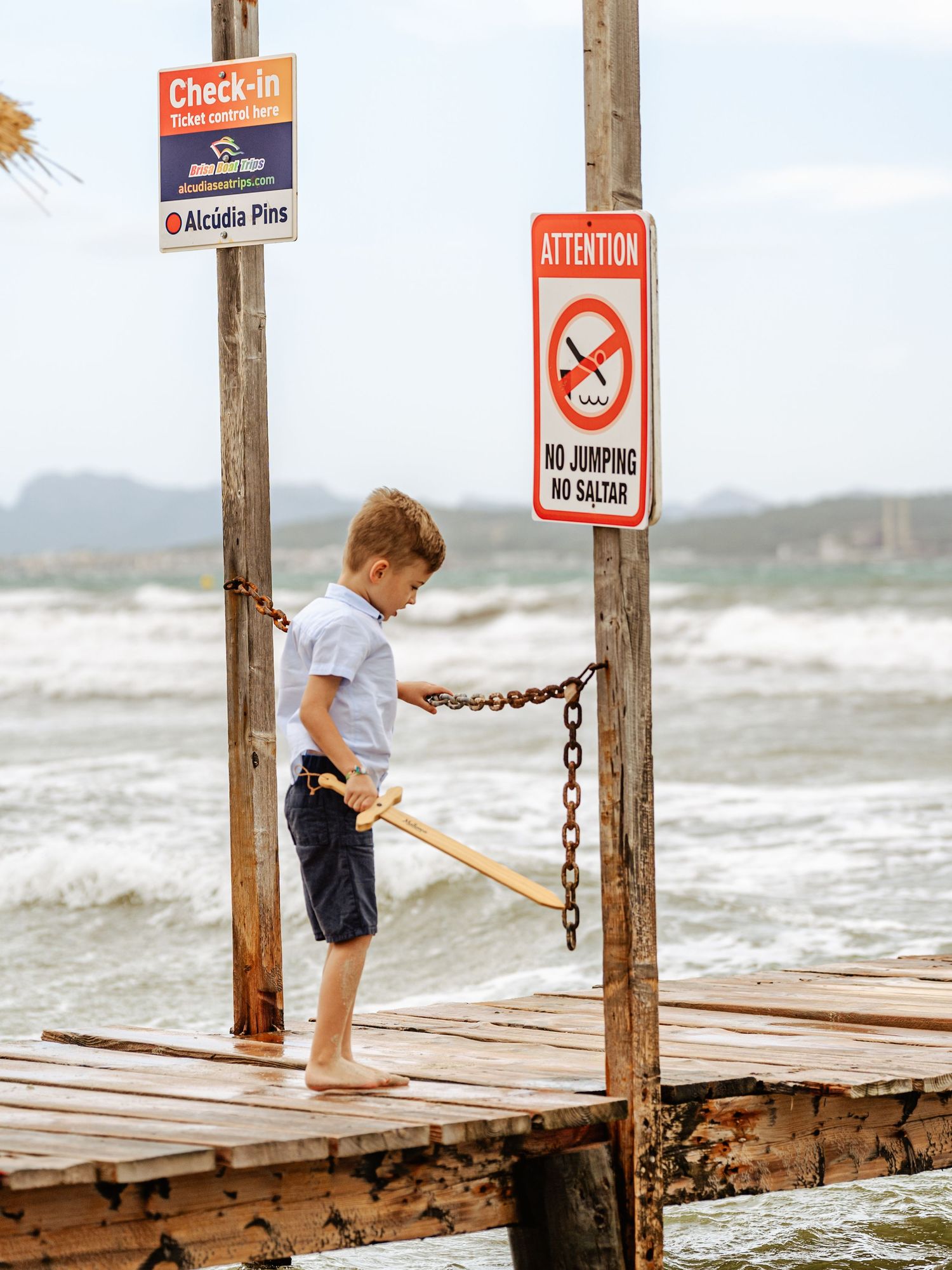 Young boy on wooden pier holding chain near no jumping sign at beach.