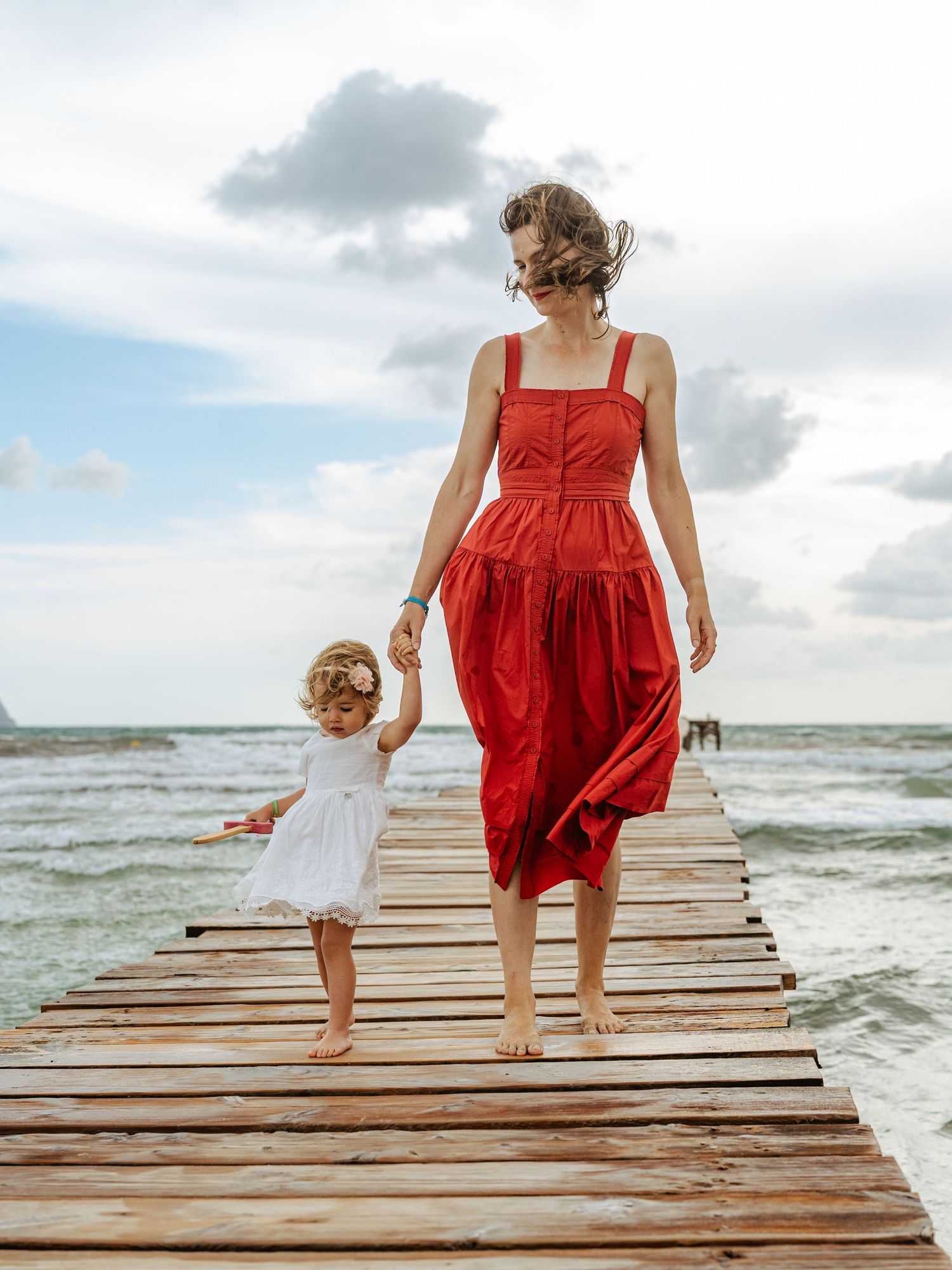Mother in red dress holding hands with toddler daughter in white dress walking on wooden beach pier.