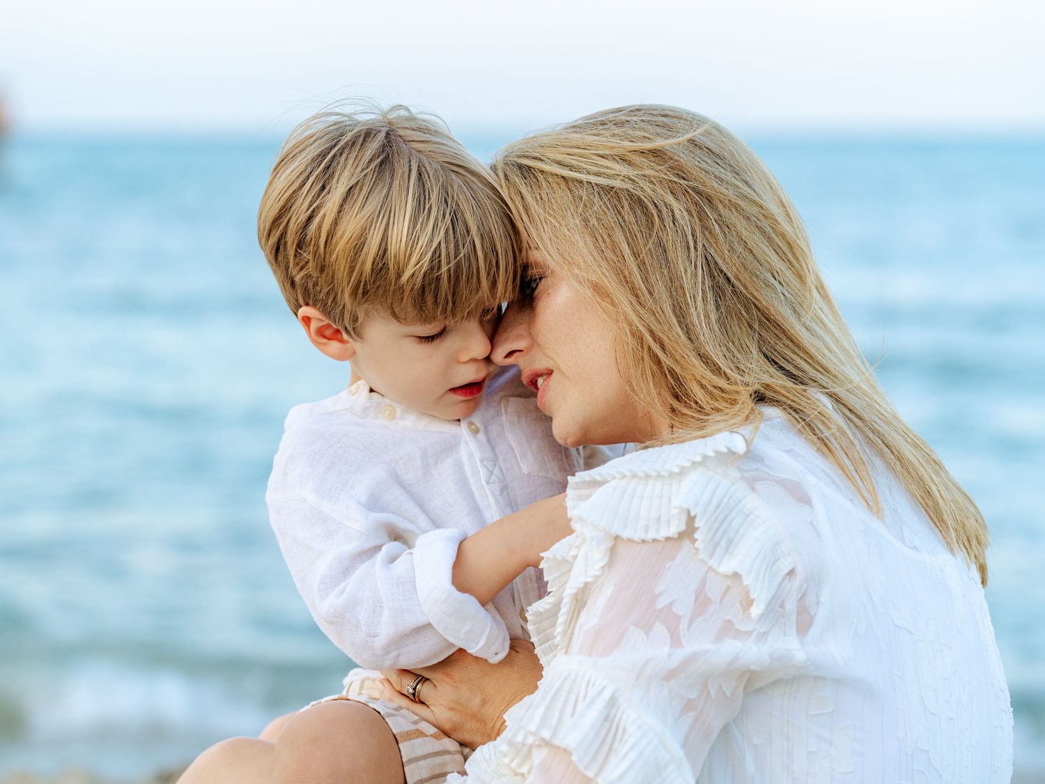 Tender moment between a blonde mother and her young son touching foreheads near the ocean shore.