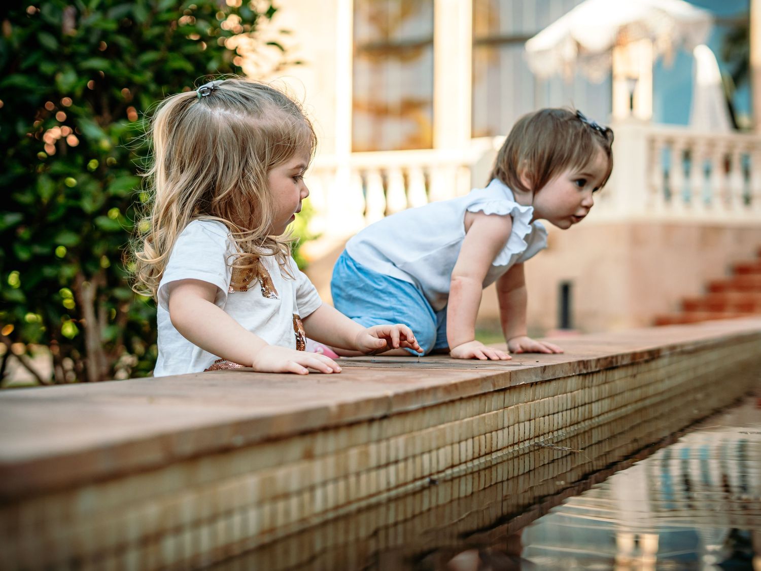 Two young girls curiously leaning over a garden water feature on a sunny day.