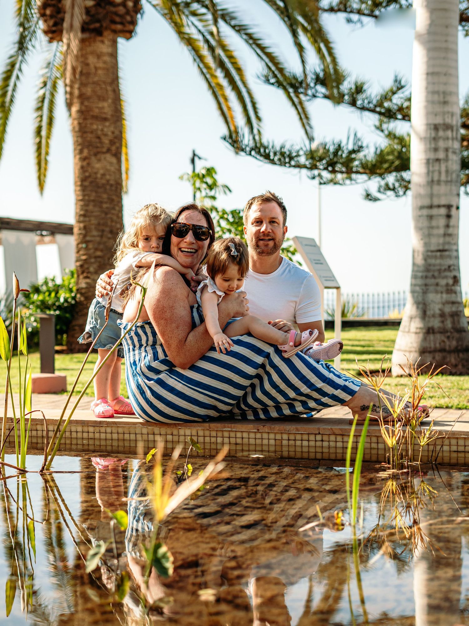 Laughing family with two young children seated by a reflective pond under palm trees.