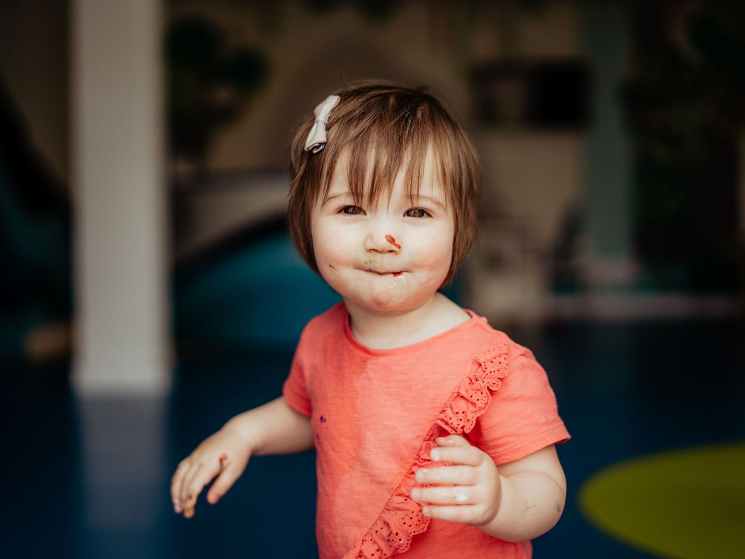 Adorable toddler girl in orange top making a funny face with hair bow accessory.