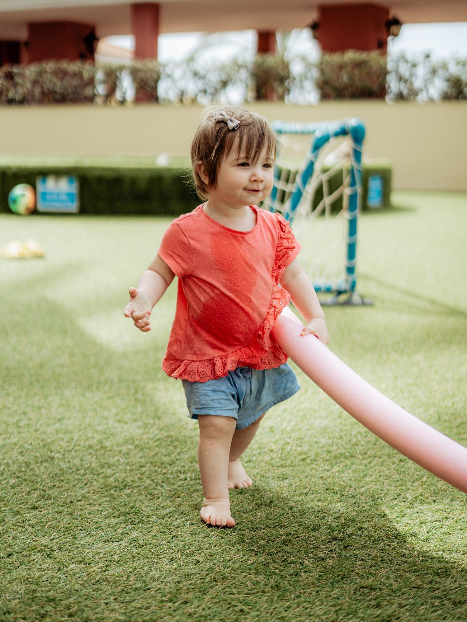 Happy toddler girl in red top walking barefoot on green grass holding a pink pool noodle.
