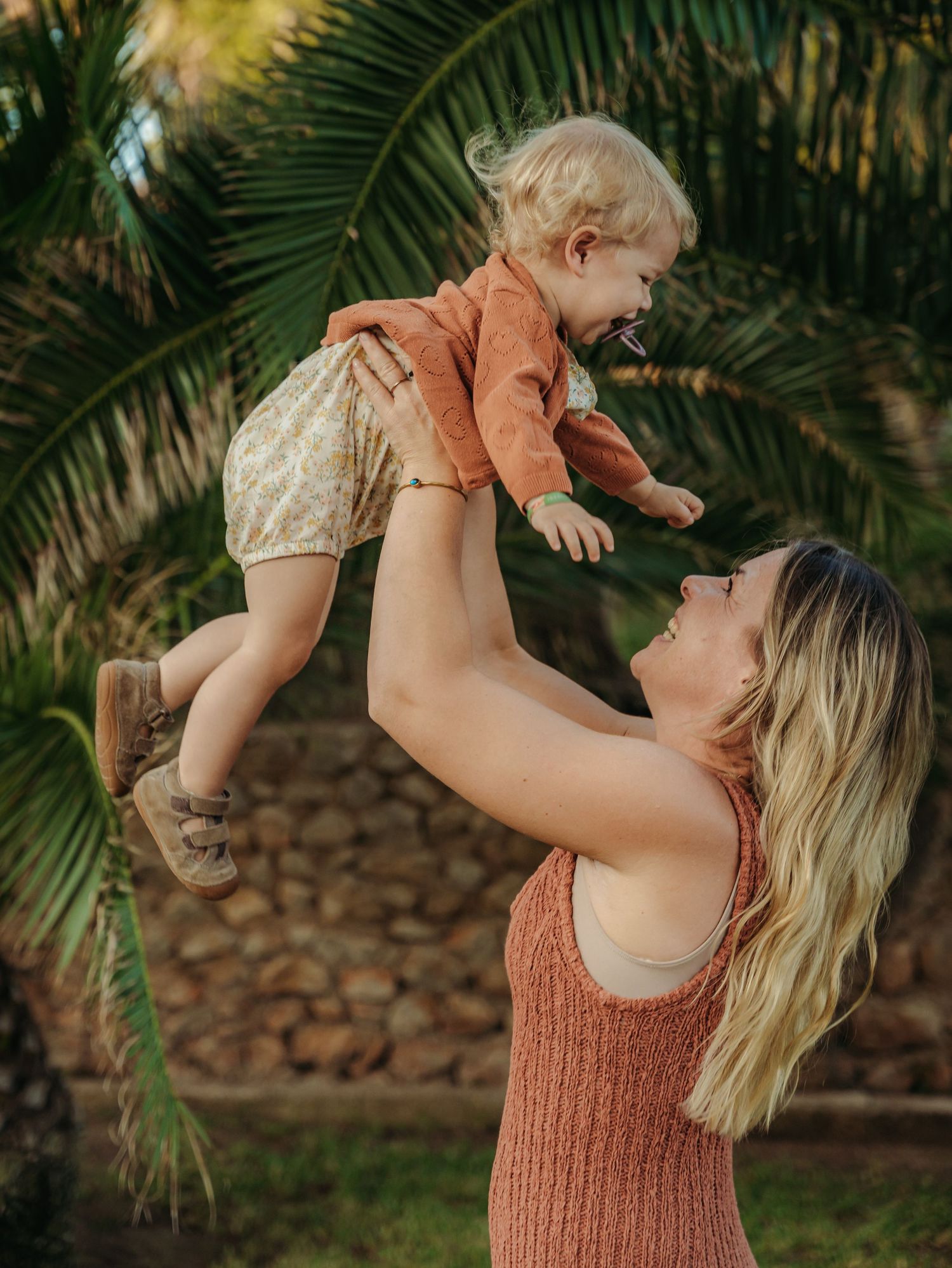 Happy mother in orange dress lifting laughing baby outdoors near stone wall and palms.