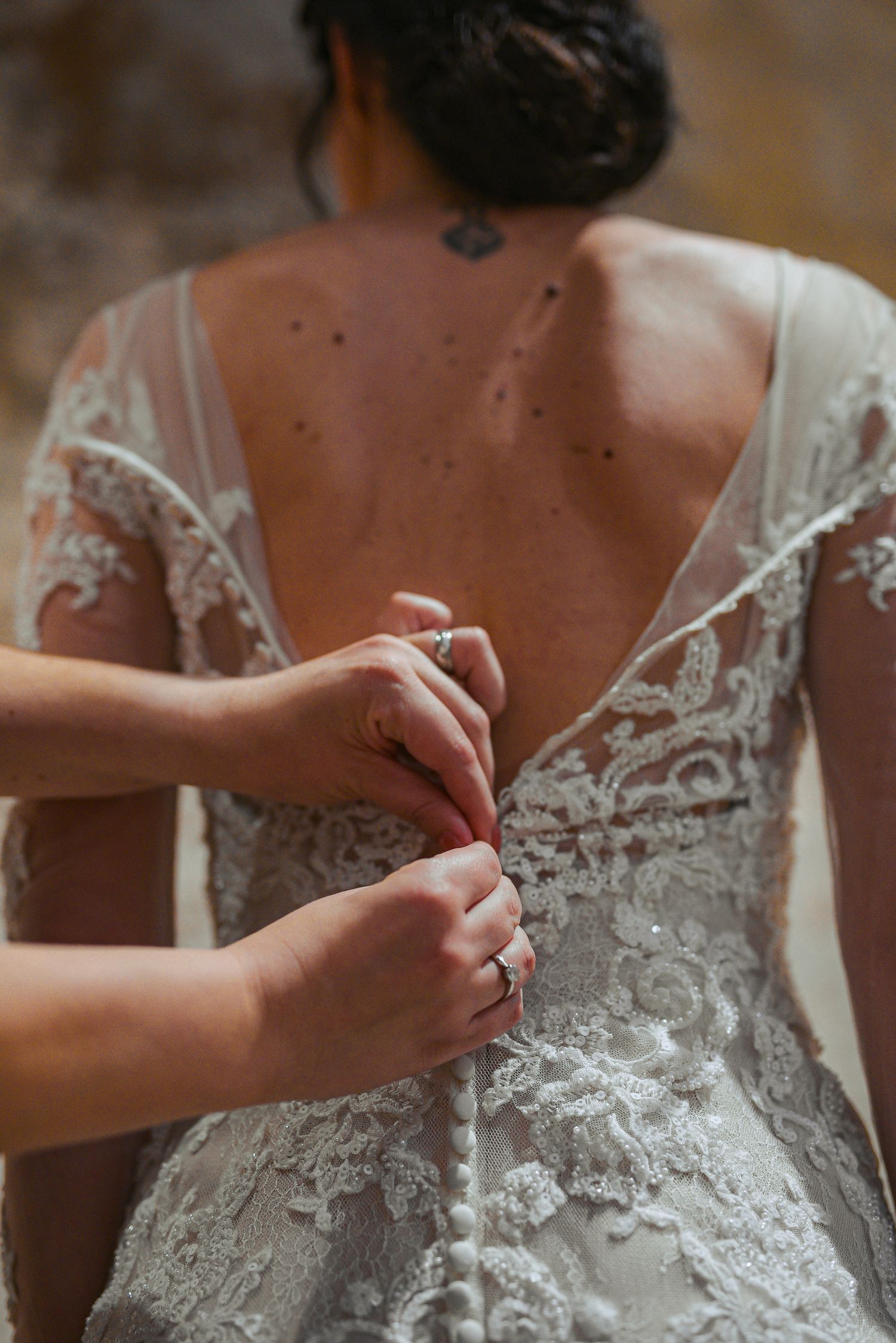 Close-up of hands buttoning a low-back lace bridal gown on a bride with a neck tattoo.