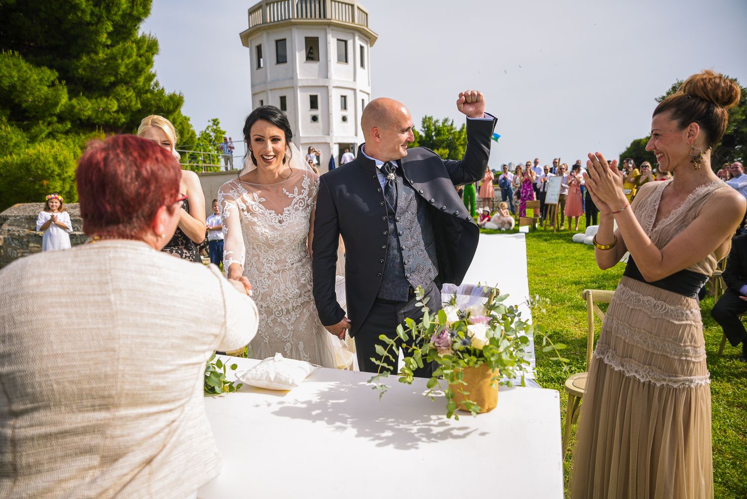 Joyful groom raises fist in celebration as bride and guests applaud at outdoor lighthouse wedding ceremony.